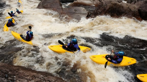 Alumnos de la Ballingarry Presentation School haciendo rafting en aguas bravas a través de un río espumoso en un entorno rocoso.