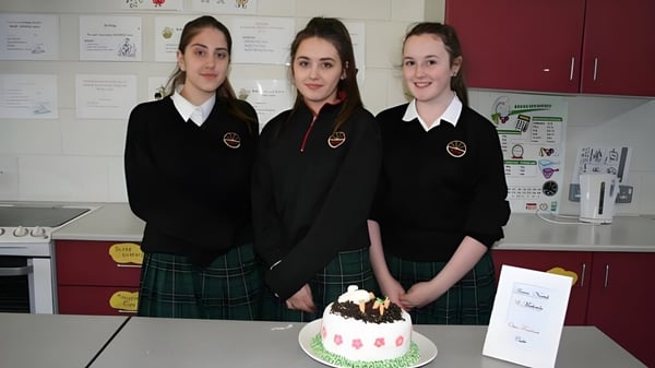 Tres estudiantes de la Ballinteer Community School están juntas en el aula frente a un pastel de cumpleaños en la mesa.