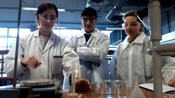 Tres personas en batas de laboratorio están en el laboratorio de la Ballinteer Community School alrededor de equipos científicos.