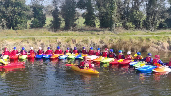 Estudiantes de la Ballymahon Vocational School reman en kayaks de colores en un lago en el área forestal.