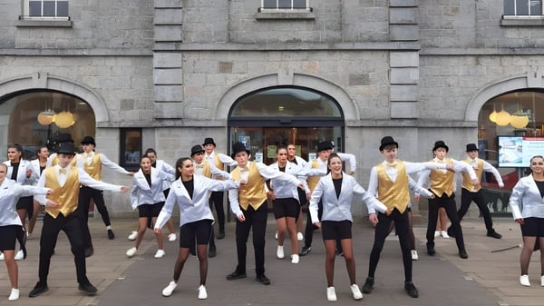 Un grupo de estudiantes de la Ballymahon Vocational School realiza una rutina de baile sincronizado en uniformes blancos y dorados frente a un edificio de piedra.