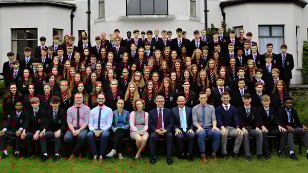 Un gran grupo de alumnas y alumnos está de pie en ropa formal frente a un edificio de la Bandon Grammar School con arquitectura clásica.