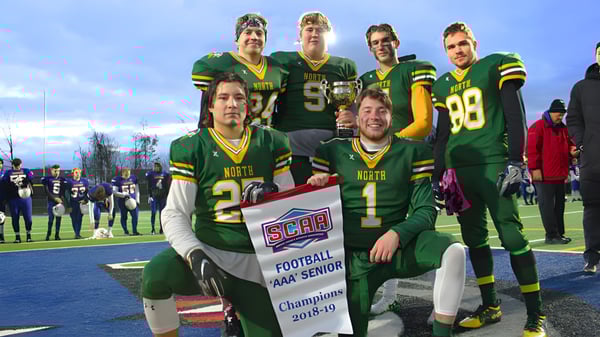 Los futbolistas del Barrie North Collegiate Institute sostienen un banner de campeonato en el campo frente a los espectadores.