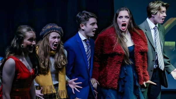 Un grupo de estudiantes de la Battle Abbey School presenta una obra de teatro en un escenario con fondo oscuro.