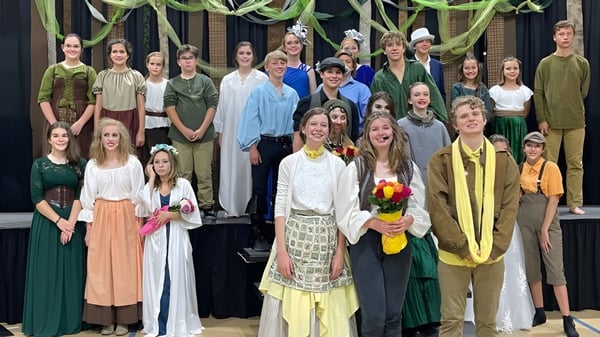 Un grupo de jóvenes estudiantes de la Bayshore Christian School está en un escenario frente a un fondo oscuro con decoraciones.