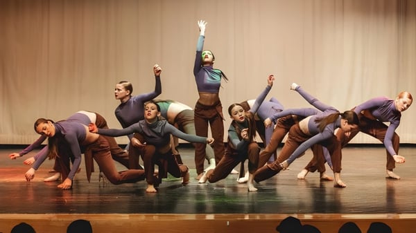 Un grupo de bailarines se presenta en el escenario de la Bear Creek Secondary School con los brazos en alto frente a un fondo de madera.