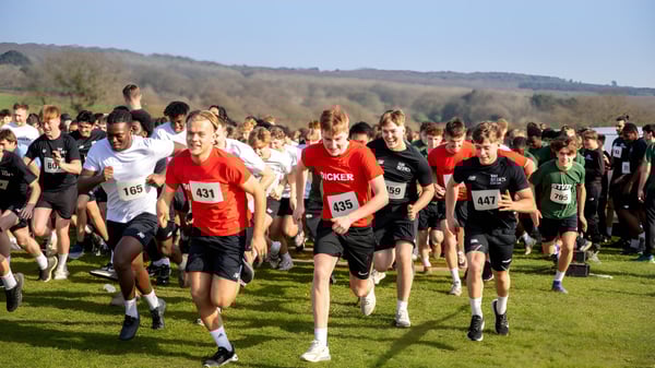 Un gran grupo de corredores participa en una carrera en una pradera con el fondo de colinas y árboles en el terreno de la Bede's Senior School.