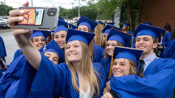 Un grupo de graduados de la Bedford School posan en togas azules para un selfie frente al campus.