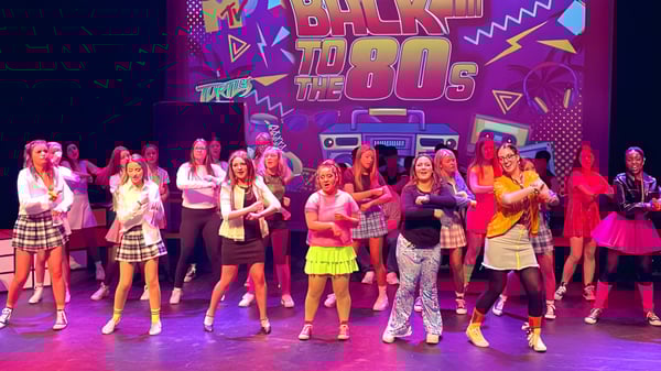 Un grupo de alumnas del Beech Hill College está en coloridos atuendos en el escenario frente a un fondo retro de 'Back to the 80s'.