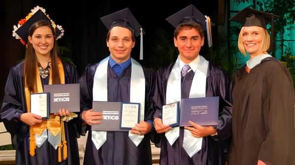 Cuatro graduados en toga de graduación en el campus del Beech Hill College sostienen sus diplomas al aire libre.
