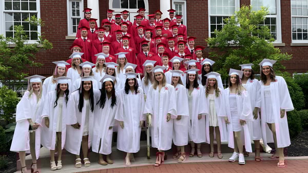 Un gran grupo de estudiantes de la Beechwood Sacred Heart School está en togas rojas y blancas frente a un edificio de ladrillo.