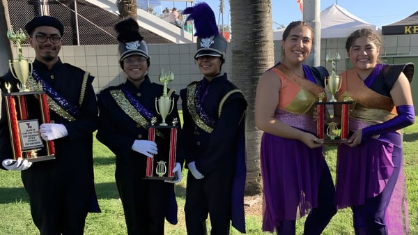 Estudiantes de la Bell High School en coloridos disfraces sostienen sus trofeos frente a un festival con carpas y palmeras.