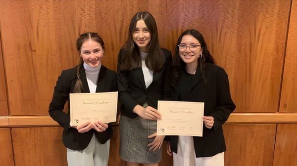 Tres mujeres en ropa formal sostienen certificados frente a un panel de madera en el campus de la Bellarmine Preparatory School.