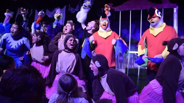 Alumnos del Belmont City College presentan una obra de teatro en un escenario con iluminación dramática.