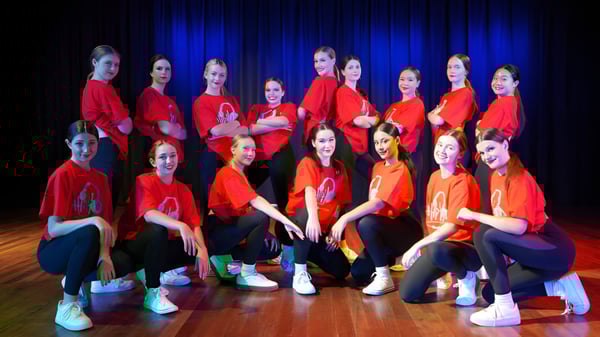 Un grupo de jóvenes estudiantes se sienta en el escenario de la Benowa State High School frente a una cortina azul.