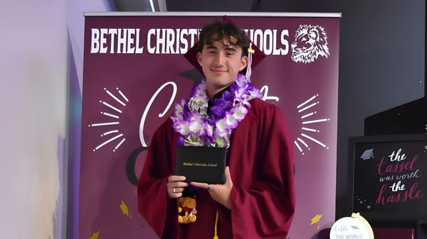 Una persona en una vestimenta roja con una corona de flores está frente a un banner morado de las Bethel Christian Schools con el logo de un león.