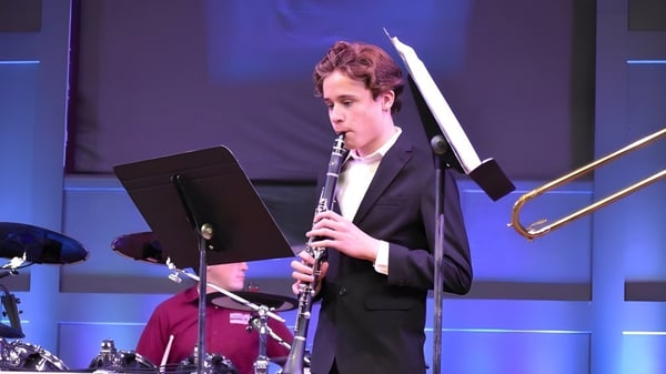 Una persona en traje toca un instrumento musical en el escenario de las Bethel Christian Schools con un fondo iluminado en azul.