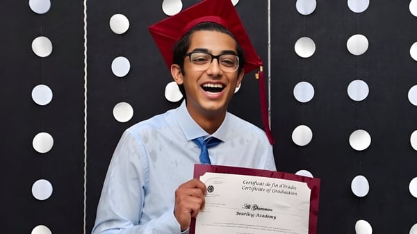 Un graduado de la Beurling Academy sostiene un diploma en su toga y birrete frente a un fondo de puntos en blanco y negro.