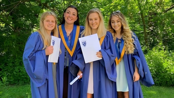 Cuatro estudiantes en togas de graduación azules están juntas en el área verde del campus del Birchmount Park Collegiate Institute.