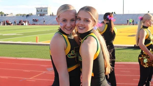 Dos animadoras de la Bishop Carroll High School posan juntas en el campo deportivo frente a un estadio.