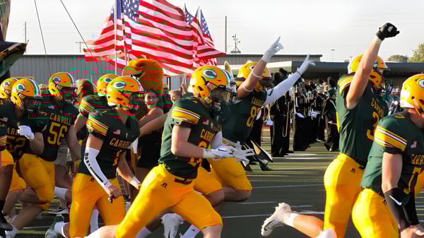Un grupo de jugadores de fútbol americano de la Bishop Carroll High School está con banderas estadounidenses en el campo de juego frente a un estadio.
