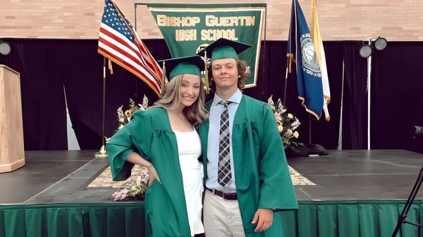 Dos estudiantes de la Bishop Guertin High School están en ropa de graduación frente a un fondo con la bandera americana y la bandera de la escuela.