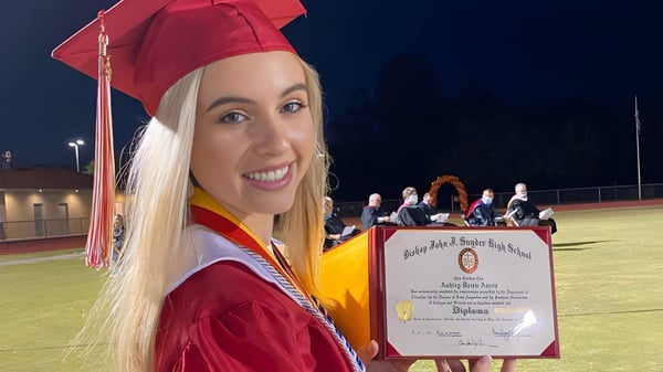 Una joven en una toga de graduación roja y birrete sostiene su diploma en el campo de fútbol de la Bishop J Snyder High School por la noche.