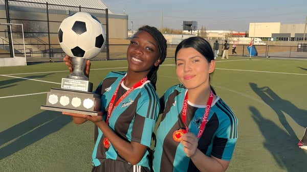 Dos estudiantes de la Bishop McNally High School sostienen un trofeo de fútbol y una pelota en el campo deportivo.