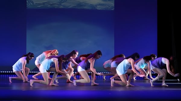 Un grupo de estudiantes de la Blackwater Community School realiza una actuación de baile sincronizada en un escenario con fondo azul.