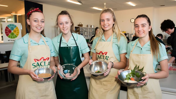 Cuatro estudiantes de la Blackwood High School llevan delantales verdes y sostienen utensilios de cocina en una cocina.