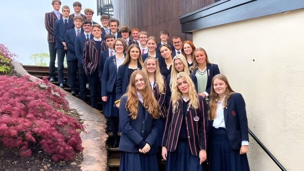 Un gran grupo de estudiantes de la Blundells School está de pie en ropa formal frente a un edificio de madera con flores en flor.