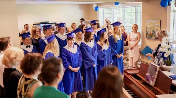 Un grupo de graduados de la Blyth Academy está junto en túnicas y gorros azules en una sala con un piano y decoración floral.
