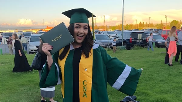 Una graduada de la Bonneville High School lleva una vestimenta de graduación verde y sostiene un diploma frente a un estacionamiento.