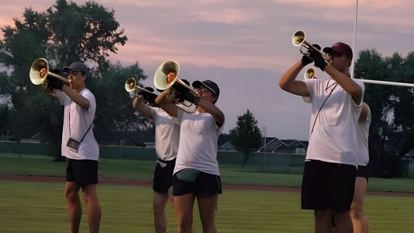 Un grupo de estudiantes de la Bonneville High School toca trompetas al aire libre frente a un campo cubierto de hierba y un cielo de atardecer colorido.
