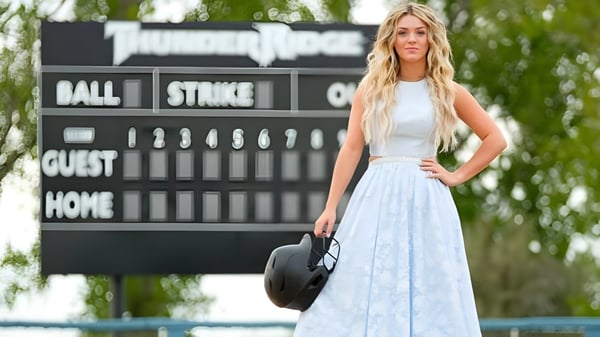 Una joven se encuentra frente al marcador de un partido de béisbol en el terreno del Bonneville Joint School District y sostiene un sombrero negro.