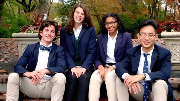 Cuatro estudiantes están sentados en un banco de piedra al aire libre en el campus de la Boston Trinity Academy frente a un fondo de árboles otoñales.