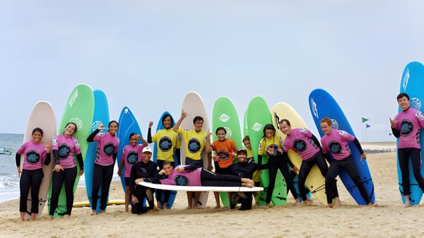 Estudiantes del Bosworth Independent College están con trajes de neopreno coloridos y tablas de surf en la playa frente al océano.