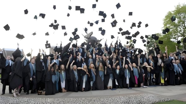 Un grupo de graduados en togas negras lanza sus gorros en el campus del Bradfield College frente a un fondo verde.