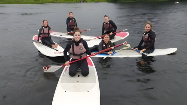 Estudiantes del Breifne College están de pie en tablas de paddle en el agua rodeados de naturaleza verde.