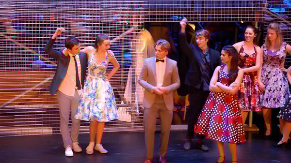 Un grupo de estudiantes de la Brentwood School está vestido con ropa formal y vestidos coloridos en un escenario con fondo de rejilla metálica.