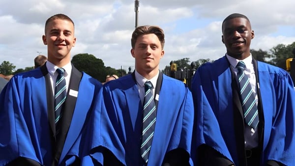 Tres estudiantes de la Brentwood School están en túnicas de graduación azules frente a un cielo nublado y árboles.