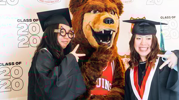 Dos estudiantes de la Britannia Secondary School en togas de graduación posan con la mascota de la escuela frente al fondo de Class of 2020.