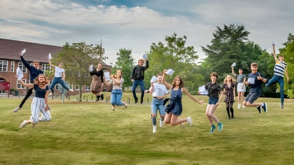 Un grupo de estudiantes del Brockenhurst College salta y celebra en un césped frente a árboles y edificios.