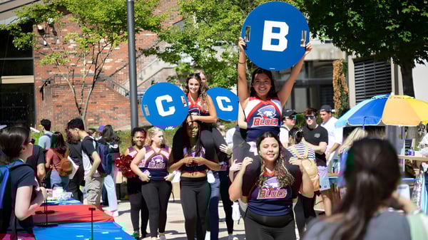 Un grupo de estudiantes sostiene carteles coloridos frente al edificio de ladrillo del Brookdale Community College en un día soleado.