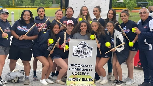 Un grupo de jóvenes estudiantes del Brookdale Community College posan con la pancarta del equipo de softball en un campo de hierba.
