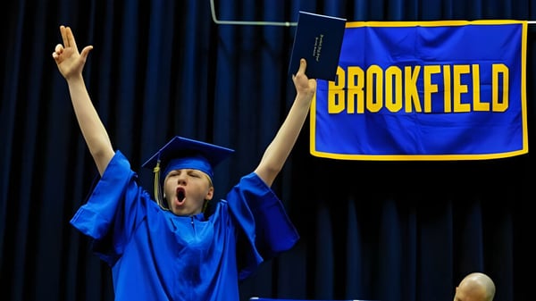 Una persona con toga de graduación azul levanta los brazos en celebración frente a un banner de la Brookfield High School.