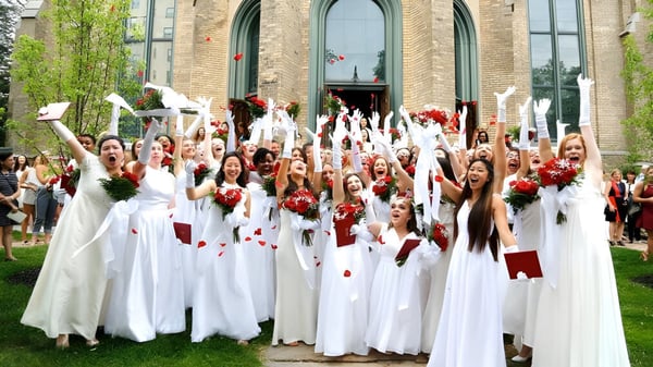 Estudiantes de Buffalo Seminary se reúnen en túnicas blancas con flores rojas frente a un edificio decorado.