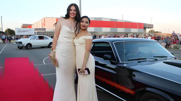 Dos mujeres en elegantes vestidos de noche posan sobre la alfombra roja frente a un edificio en el campus de la Bundaberg State High School.