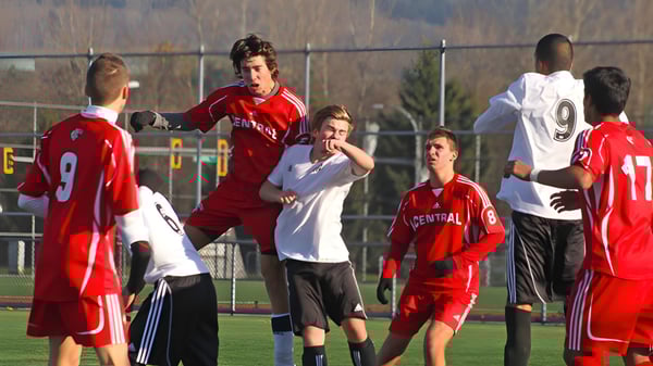 Estudiantes de la Burnaby Central School juegan al fútbol en un campo de césped frente a una cerca y árboles.