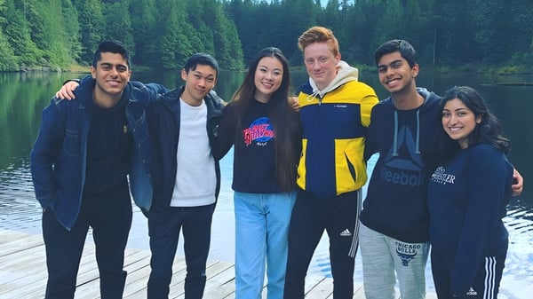 Un grupo de alumnas y alumnos de la Burnaby Mountain School está en un muelle de madera frente a un lago tranquilo con árboles verdes.